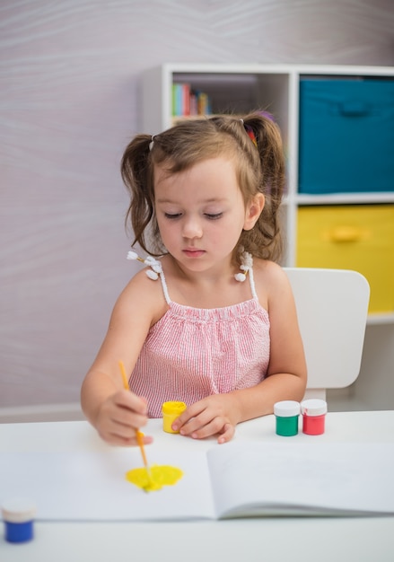 Ein kluges Mädchen mit Zöpfen sitzt am Tisch und zeichnet ein Album mit einem Pinsel mit Farben im Kinderzimmer