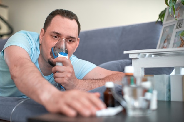 Ein kranker Mann atmet durch eine Inhalationsmaske. auf der Couch liegen und zieht seine Hand zur Heilung