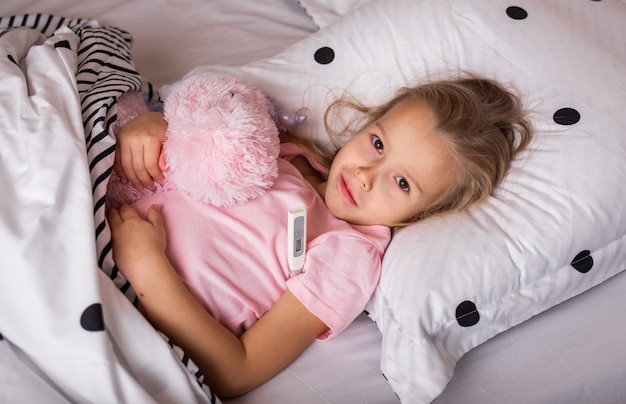 Ein krankes kleines blondes Mädchen im Schlafanzug liegt mit einem Stofftier im Schlafzimmer und misst die Temperatur mit einem Thermometer