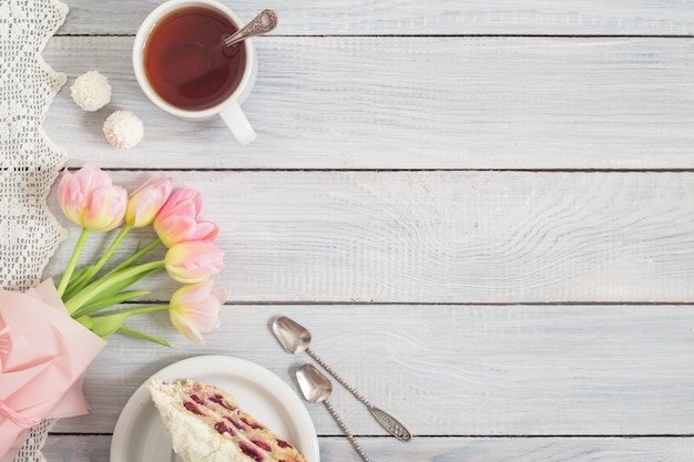 Ein Kuchen mit Kirschen, einer Tasse Tee und rosa Tulpen auf einem weißen Holztisch