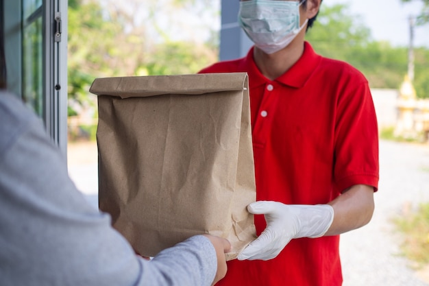 Ein Kurier, der eine rote Uniform mit Handschuhen und Gesichtsmasken trägt, um das Risiko einer Kontamination bei der Lieferung von Lebensmitteln in die Hände der Kunden zu verhindern. Lebensmittellieferdienste