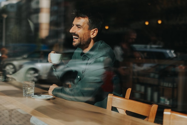 Ein lachender Mann genießt eine warme Tasse Kaffee, während er in einem sonnigen Café sitzt und sich in einer fröhlichen Nachmittagsatmosphäre mit Blick auf die Stadt sonnt