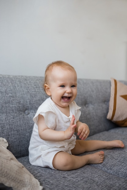 Ein lachendes Baby in einem Musselin-Overall sitzt auf dem Sofa im Wohnzimmer