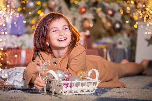 Ein lachendes Mädchen liegt mit einer Neujahrsdekoration in den Händen vor dem Hintergrund eines Weihnachtsbaumes auf dem Boden.