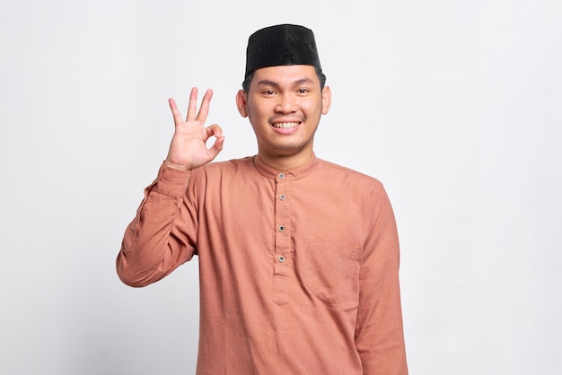Ein lächelnder asiatischer muslimischer Mann, der eine Okay-Geste zeigt, zeigt ein Symbol der Zustimmung, das über weißem Hintergrund isoliert ist