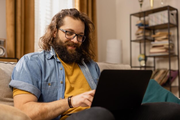 Ein lächelnder bärtiger Mann mit Brille arbeitet fern von zu Hause aus, der Mann benutzt einen Laptop, um seine Favoriten zu durchsuchen