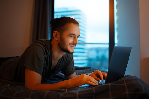 Ein lächelnder junger Mann, der im Bett liegt und zu Hause am Laptop arbeitet, auf der Tastatur tippt