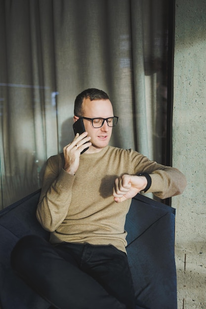 Ein lächelnder junger Mann mit Brille lächelt und telefoniert mit einem Mobiltelefon, während er sich im Büro entspannt Ein junger Freiberufler arbeitet aus der Ferne