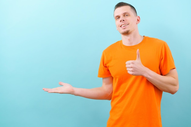 Ein lächelnder kaukasischer Mann in einem orangefarbenen T-Shirt, der eine Hand auf der unteren Seite hält und seinen Daumen nach oben zeigt. Leerer Kopierbereich auf blauem Hintergrund