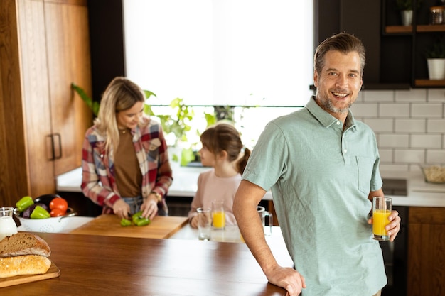 Ein lächelnder Mann hält ein Glas Orangensaft, während zwei Frauen in einer hellen Küche kochen