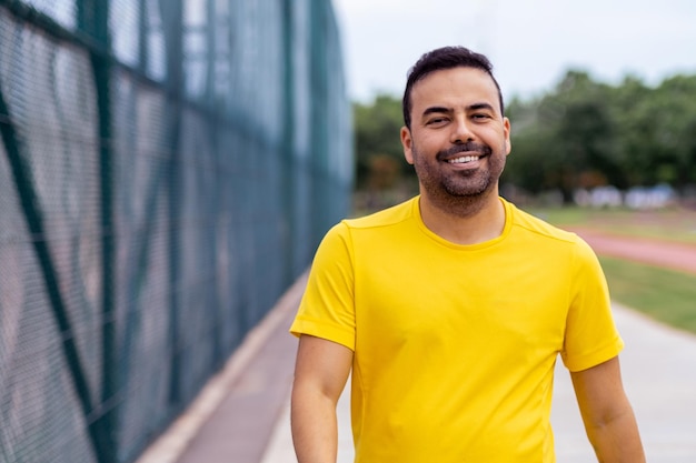 Ein lächelnder Mann in gelbem T-Shirt genießt einen Spaziergang entlang eines öffentlichen Sportplatzes im Park, um sich gesund zu fühlen