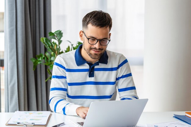 Ein lächelnder Mann mit Brille sitzt am Schreibtisch im Büro und surft auf einem Laptop im drahtlosen Internet Glücklicher männlicher Arbeiter entspannt sich in der Arbeitspause