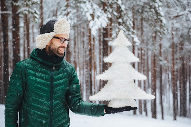 Ein lächelnder Mann mittleren Alters sieht mit glücklichem Ausdruck auf einen künstlichen weißen kleinen Tannenbaum, der die Neujahrsferien feiern will, und verbringt den Morgen bei Schneewetter an der frischen Luft im Wald