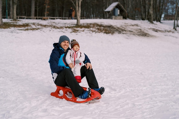 Ein lächelnder Vater und seine kleine Tochter sitzen auf einem Schlitten auf einer schneebedeckten Ebene
