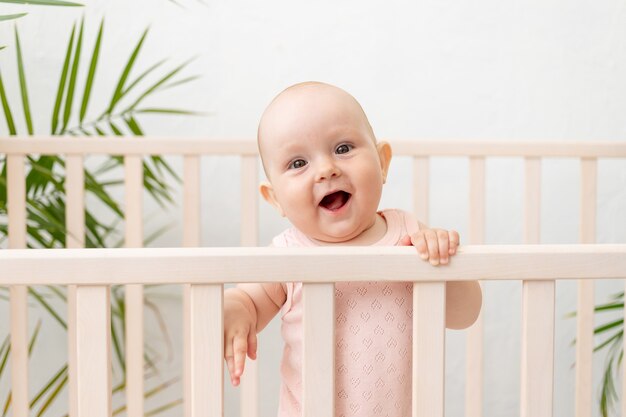 Ein lächelndes Baby in einer Krippe in einem rosa Body für sechs Monate steht an der Seite fest und lacht