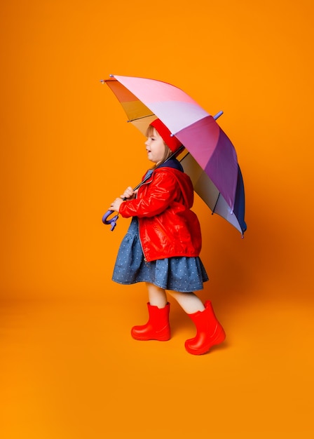 Ein lächelndes Kindermädchen in einer roten Jacke und Stiefeln, das einen Regenschirm hält, steht auf einem gelben Hintergrund. kleines Mädchen in hellen Kleidern hält einen Regenbogenregenschirm.