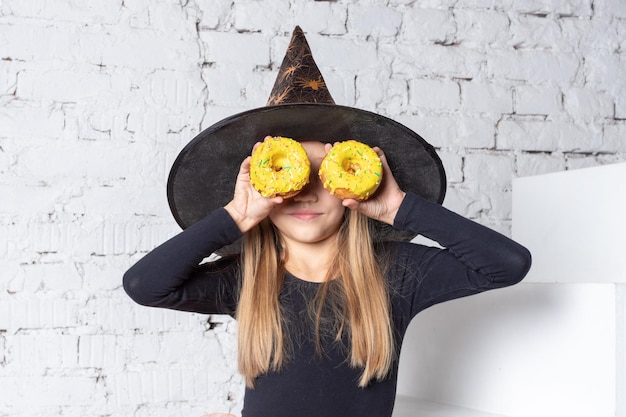 Ein lächelndes kleines blondes Mädchen in einem Hexenkostüm, das gelbe Donuts in ihren Händen hält und auf den Stufen des Hauses sitzt