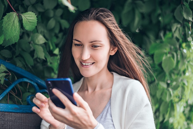 Ein lächelndes Mädchen in der weißen Jacke, die auf Bank sitzt und in Smartphone schaut