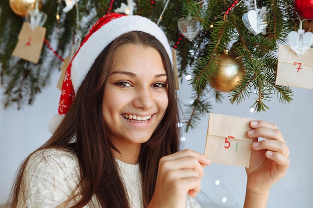 ein lächelndes Mädchen in einer Weihnachtsmütze mit einem Adventskalender diy in den Händen in der Nähe des Weihnachtsbaums