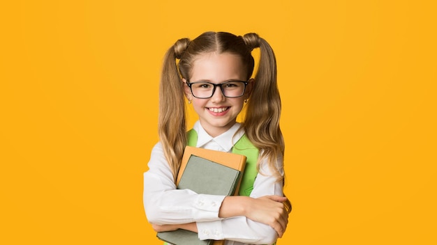 Ein lächelndes Schulkind, ein Grundschüler, ein Mädchen, das Bücher umarmt und froh ist, dass die Schule auf Gelb steht.