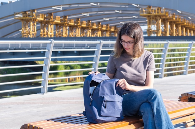 Ein lächelndes Studentenmädchen mit Brille, das ihren Rucksack öffnet und nach etwas in ihrer Tasche sucht, das draußen auf einer Bank sitzt