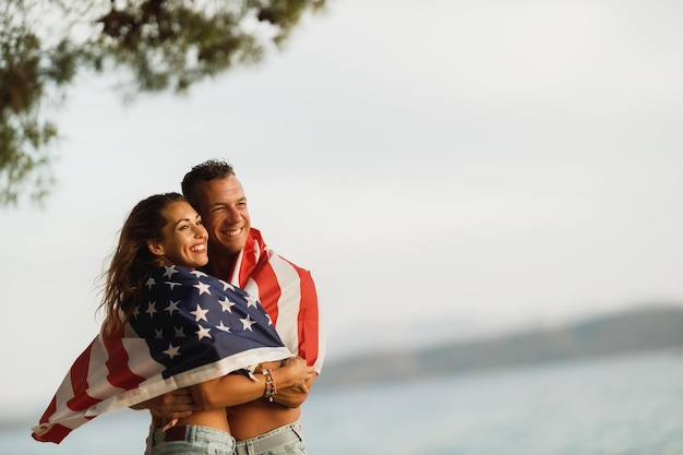 Ein lächelndes, umarmtes Paar mit US-Nationalflagge genießt einen entspannten Tag am Strand.