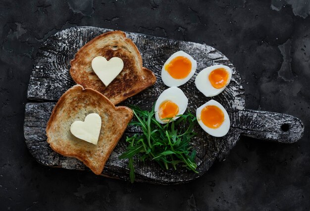 Ein leckeres, einfaches Frühstück, gekochte Eier mit weichem Eigelb, Arugula-Toast mit Butter auf einem rustikalen Schneidbrett auf dunklem Hintergrund