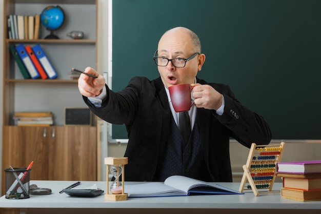 Ein Lehrer mit Brille sitzt an der Schulbank vor der Tafel im Klassenzimmer und hält eine Tasse Kaffee in der Hand, die mit einem Bleistift auf etwas zeigt, das überrascht wird