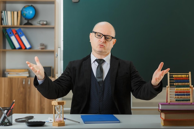 Ein Lehrer mit Brille und Klassenbuch sitzt an der Schulbank vor der Tafel im Klassenzimmer und blickt in die Kamera, die unzufrieden die Arme in Unmut und Empörung hebt