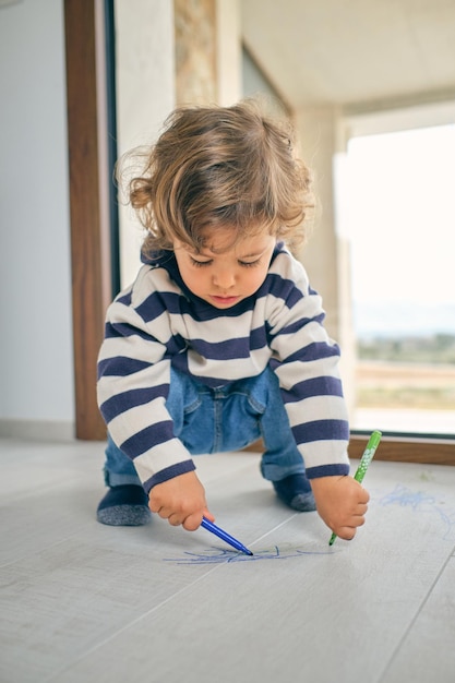 Ein liebenswertes, fokussiertes, lockighaariges Kind in lässiger Kleidung, das auf Hüften sitzt und auf dem Laminatboden mit farbenfrohen Markierungen in der Nähe des Fensters im Zimmer krabbelt