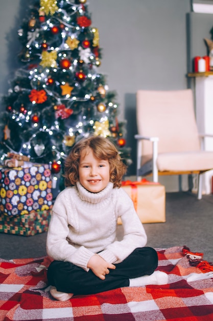 Ein lockiger Junge von zehn Jahren ohne Vorderzähne sitzt mit Geschenken in einem Pullover am Neujahrsbaum, schaut in den Rahmen und lächelt.