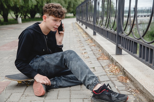 Ein lockiger Teenager mit europäischem Aussehen in schwarzem Hoodie sitzt auf einem Skateboard in einer Gasse und k...