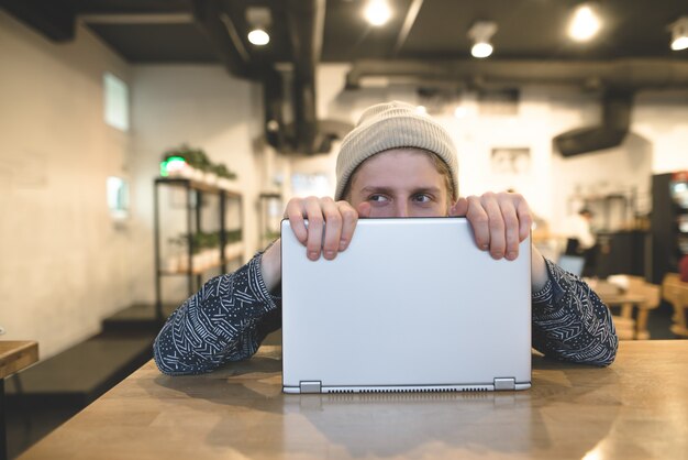Ein lustiger junger Mann, der sich hinter einem Laptop versteckt. Fröhliche Hipster arbeiten am Computer in einem gemütlichen Café. Schau weg.