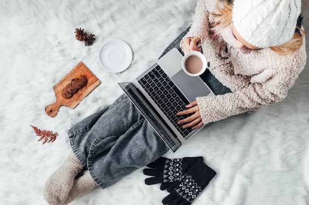 Ein Mädchen, das zu Hause mit Laptop arbeitet und die Wintersaison mit heißer Schokolade und Keksen genießt
