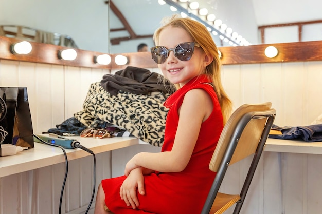 Ein Mädchen, ein Kind, ein Modell in einem schönen Kleid und einer Sonnenbrille, das in der Nähe eines Spiegels mit Lampen und Lächeln sitzt