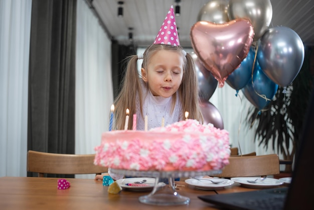 Ein Mädchen feiert seinen Geburtstag über das Internet zur Quarantänezeit bläst die Kerzen auf der Geburtstagstorte in der Nähe des Laptops aus Festliche Dekoration Süßigkeiten Luftballons Töchter mit Eltern Kinderfest