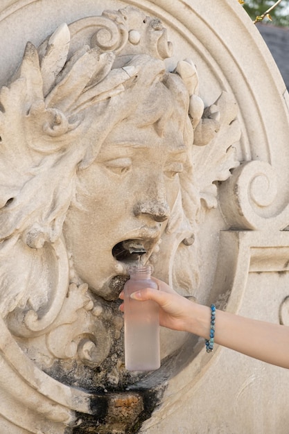 Ein Mädchen gießt aus einem Trinkbrunnen in Form eines menschlichen Gesichts in Österreich Wasser in eine Flasche