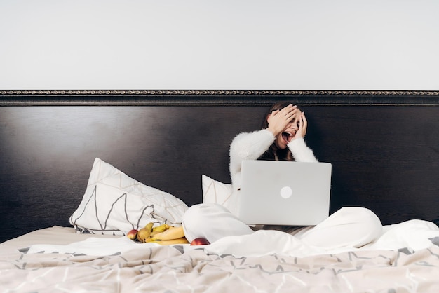 Ein Mädchen im weißen Schlafanzug sitzt im Bett und sieht sich auf ihrem Laptop einen Gruselfilm an, der ihr Gesicht mit den Händen bedeckt