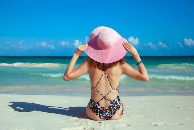 Ein Mädchen in Bikinis und rosa Hut, Foto von hinten. Junge Dame, die am Strand ein Sonnenbad nimmt. Schöne Frau, die am Sommersandstrand aufwirft. Sommerporträt im Freien der hübschen Sportartfrau im Hut.