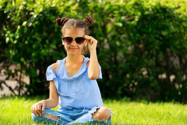 Ein Mädchen in blauer Kleidung sitzt im Sommer im Park auf dem Gras. ein Kind mit Sonnenbrille. Foto in hoher Qualität