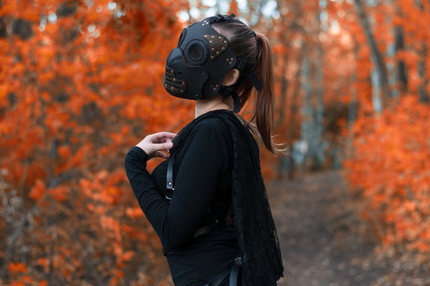Ein Mädchen in einem BDSM-Kostüm und einer schwarzen Maske in einem roten Wald. Eine Idee für Halloween.