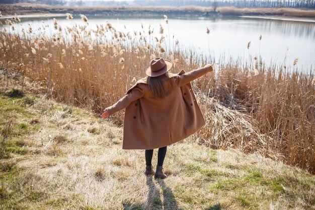 Ein Mädchen in einem braunen Mantel, Hut und Brille geht in einem Park mit einem See unter der hellen Sonne spazieren. Erfreut sich am Leben und lächelt. Der Frühlingsanfang