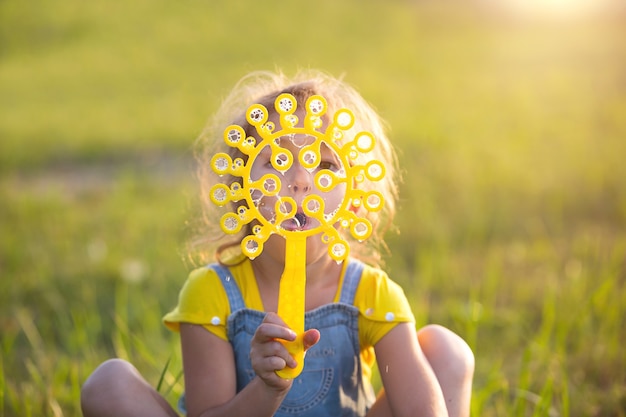 Ein Mädchen in einem Denim-Overall bläst im Sommer auf einem Feld bei Sonnenuntergang Seifenblasen. Internationaler Kindertag, glückliches Kind, Outdoor-Aktivitäten. Sommer Hintergrund. Gesunder und umweltfreundlicher Lebensstil