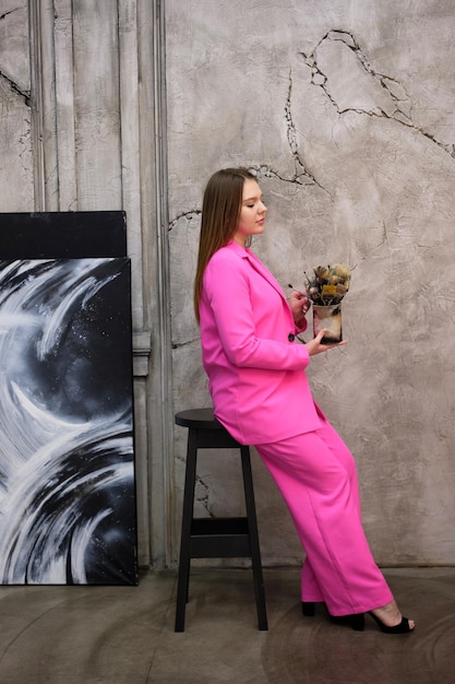 Ein Mädchen in einem formellen Anzug sitzt auf einem Barhocker mit einem Glas Pinsel vor dem Hintergrund einer strukturierten Wand neben einem Gemälde