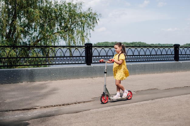 Ein Mädchen in einem gelben Kleid lernt, einen zum Geburtstag geschenkten Roller entlang des Stadtsees zu fahren