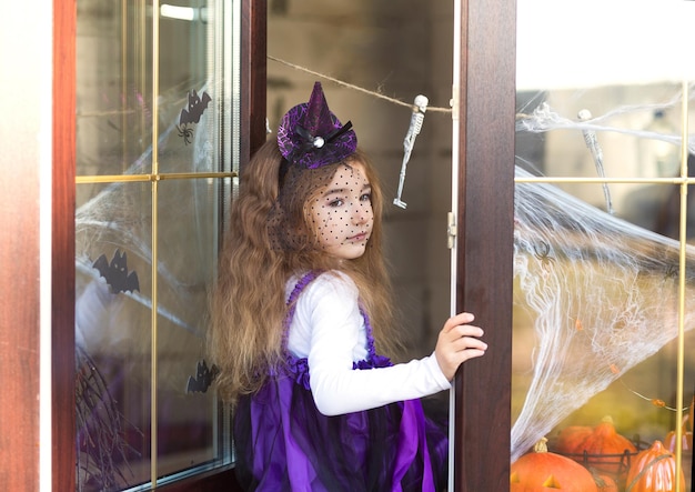 Ein Mädchen in einem Hexenkostüm auf der Fensterbank am Fenster bei einer Halloween-Party, die das Haus schmückt Spinnweben Fledermäuse Spinnen Kürbisse Schreckliche Landschaft Angst und Schrecken