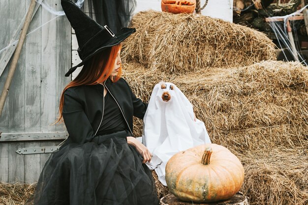 Ein Mädchen in einem Hexenkostüm mit einem Hund in einem Geisterkostüm hat Spaß auf der Veranda eines Hauses, das dekoriert ist, um eine Halloween-Party zu feiern