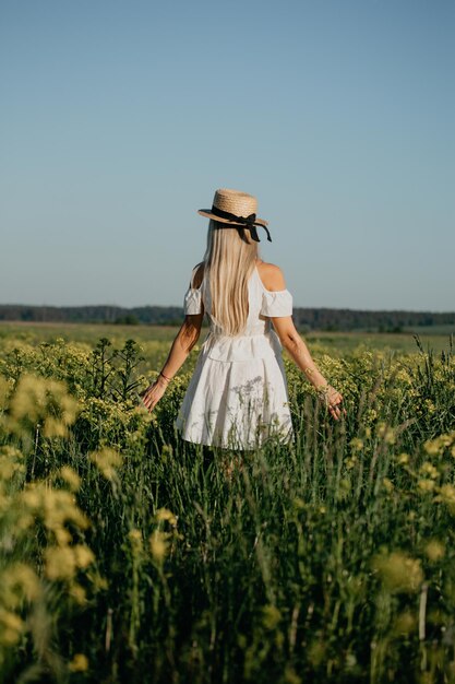 Foto ein mädchen in einem hut und einem weißen kleid steht mit dem rücken zu einem blühenden gelben raps