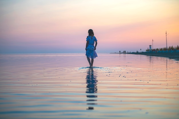 Ein Mädchen in einem Kleid am Strand bei Sonnenuntergang geht auf dem Wasser spazieren