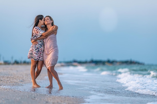 Ein Mädchen in einem Kleid umarmt ihre Mutter in einem Kleid, während sie am Strand in der Nähe des Meeres mit Wellen entlang geht